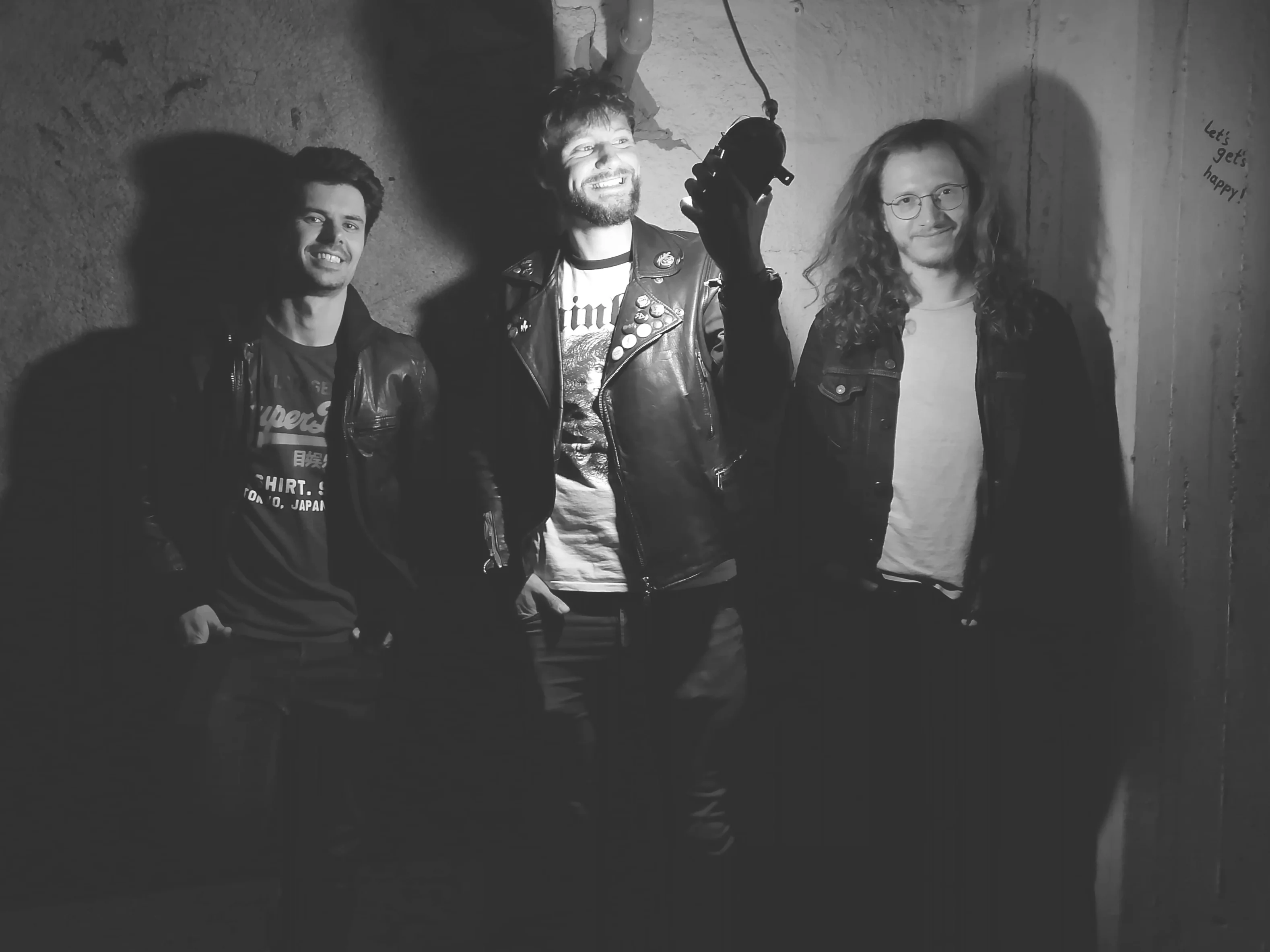 Black and white bandphoto of Bad Age.
Three guys standing in front of a bare wall. Lighted by a cellar lamp the middle one is holding in his hand, facing it.