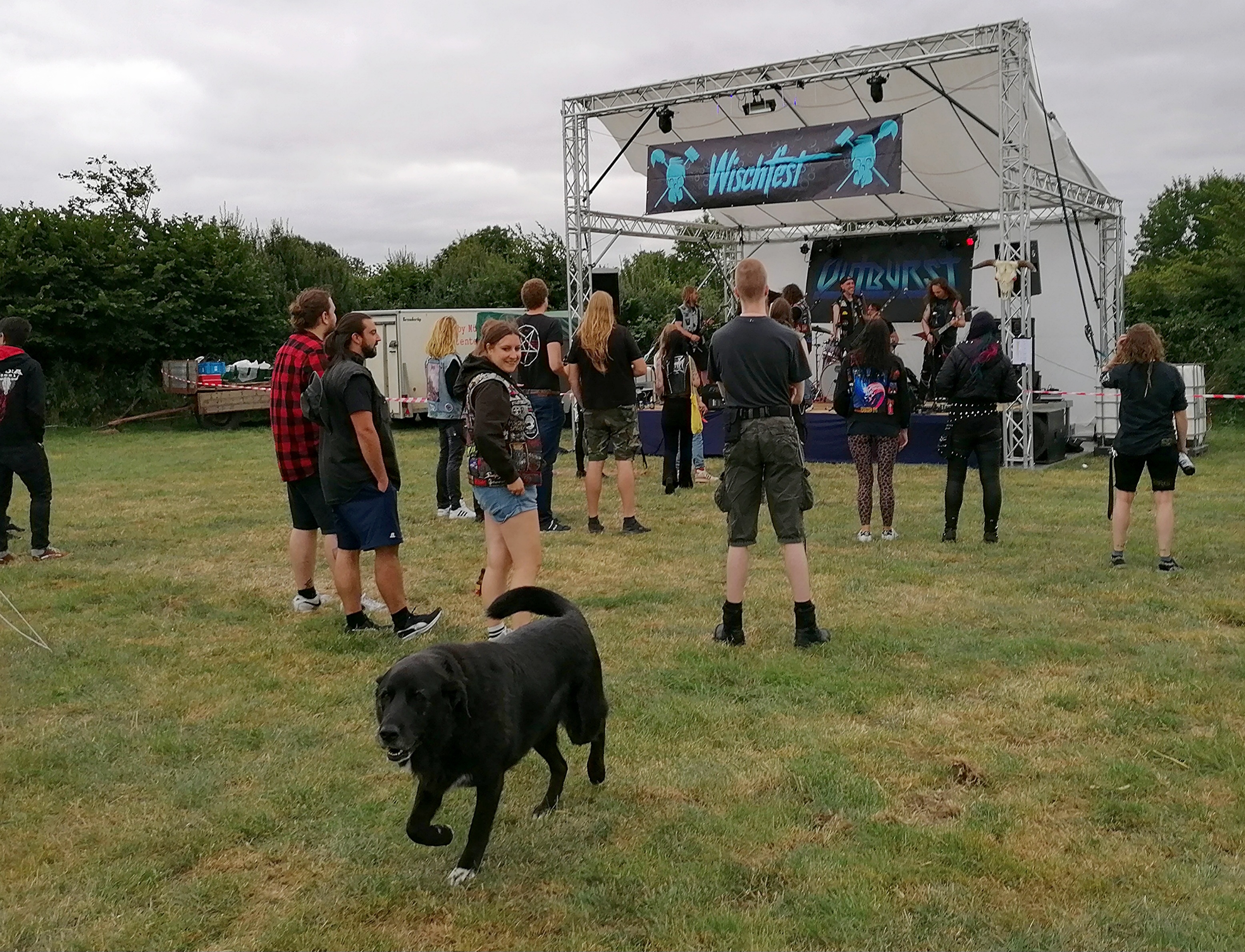 The farm dog Balu, a big black dog, runs past the camera across the grass. Behind him is the stage, guests are watching a band play.