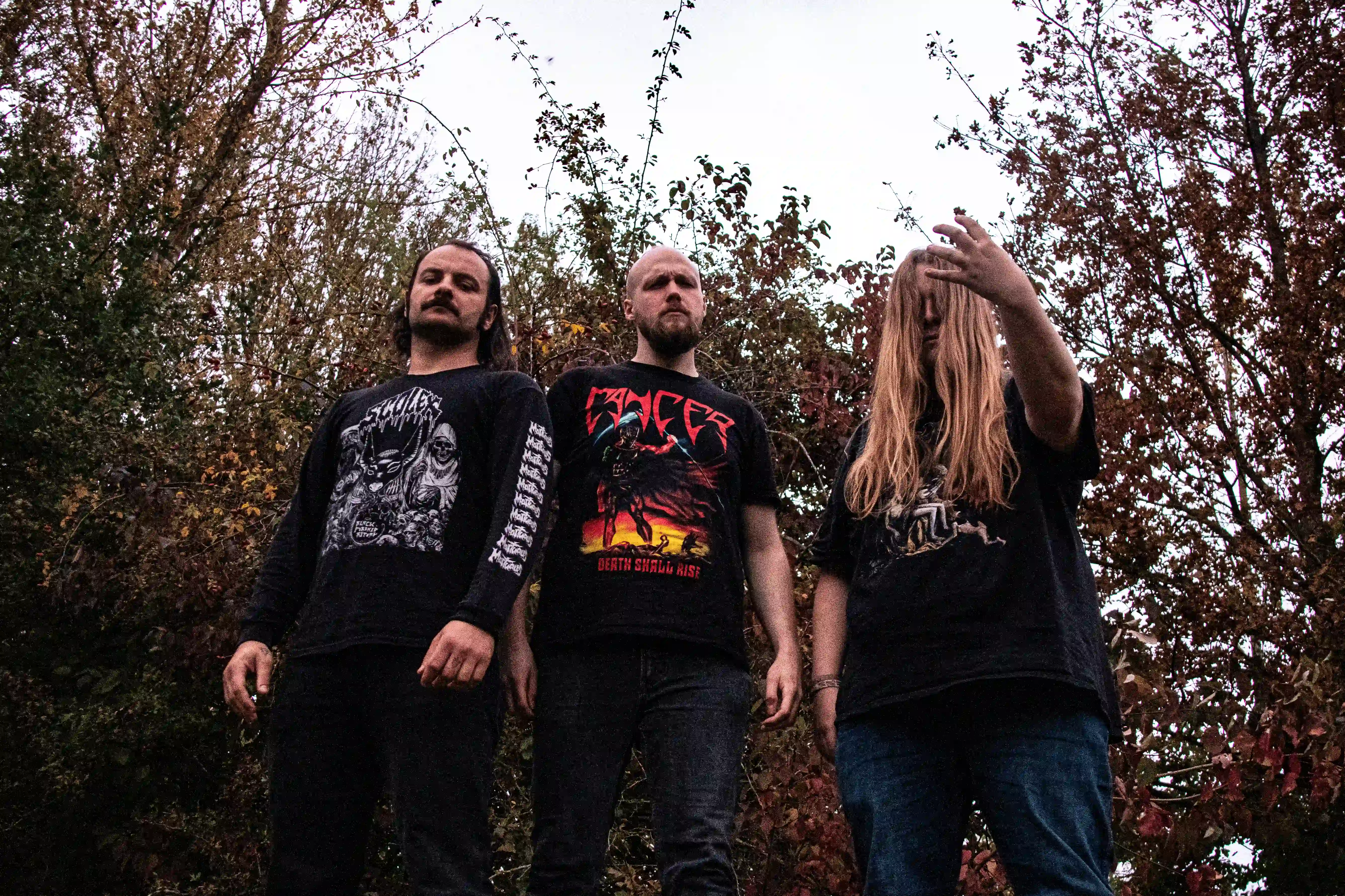 Bandfoto von Monstrum.
Das Bild ist von unten fotografiert. Man sieht die drei Männer von den Kien aufwärts. Hinter ihnen sind Bäume mit verschiedenfarbigem Laub und ein weißer Himmel.
Die zwei linken Männer schauen in die Kamera. Der Mann rechts hat seine langen blonden Haare vor dem Gesicht. Seine Augen sind nicht zu sehen. Er hat die linke Hand in epischer Pose gehoben.