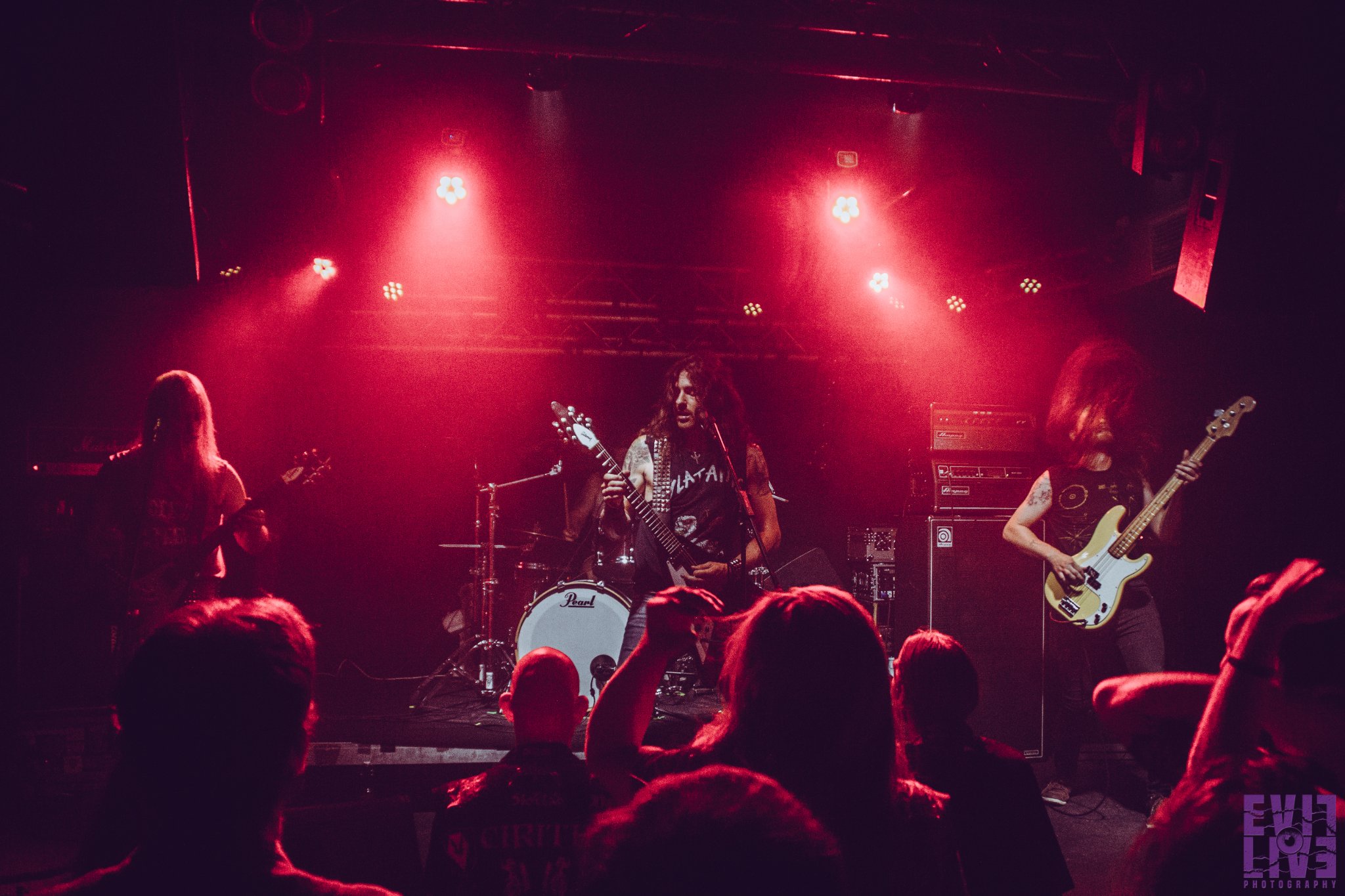 Bandfoto von Blitz.
Livefoto von einem Auftritt. Die Bühne ist in rotes Licht getaucht. Drei langhaarige Männer im Vordergrund, im Hintergrund erkennt man Schlagzeug und Drummer nur zum Teil.
Links Gitarre, mitte Gitarre, rechts Bass.
Im Vordergrund seht das Publikum mit dem Rücken zur Kamera.