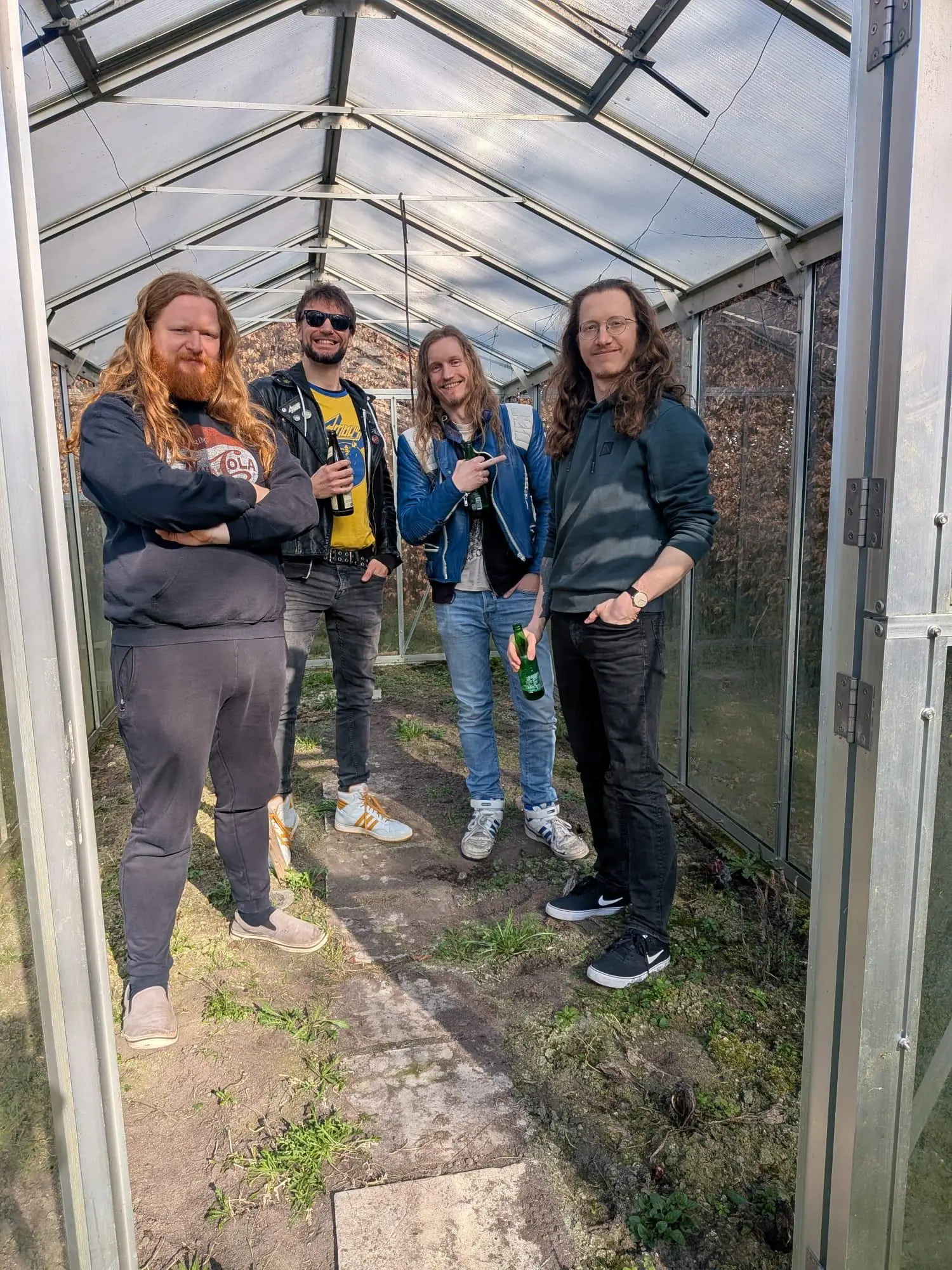 Three men are standing in a greenhouse, looking into the camera.
The two in the middle are grinning, one of them subtly holding up his middle finger.
