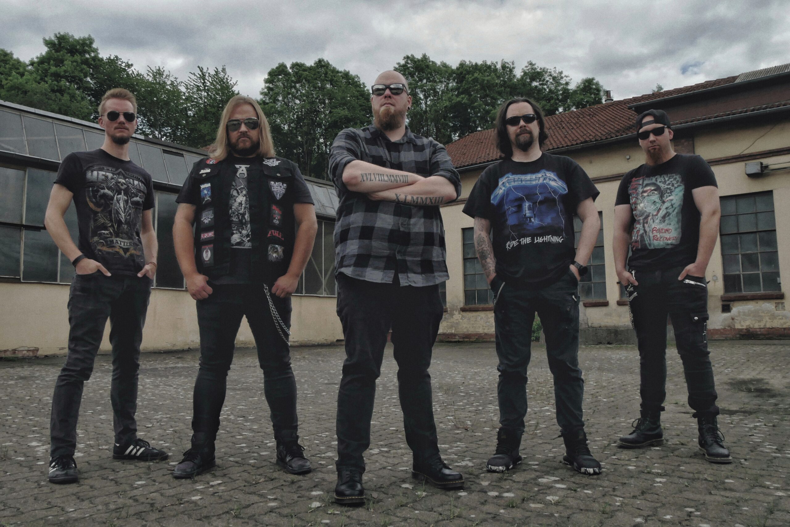 Bandphoto of Erasement. Five men, mostly dressed in black and wearing sunglasses, are standing in a row at some distance in front of abandoned-looking buildings. In the background you can see trees towering over the buildings and a cloudy sky. The photo is taken slightly from below.