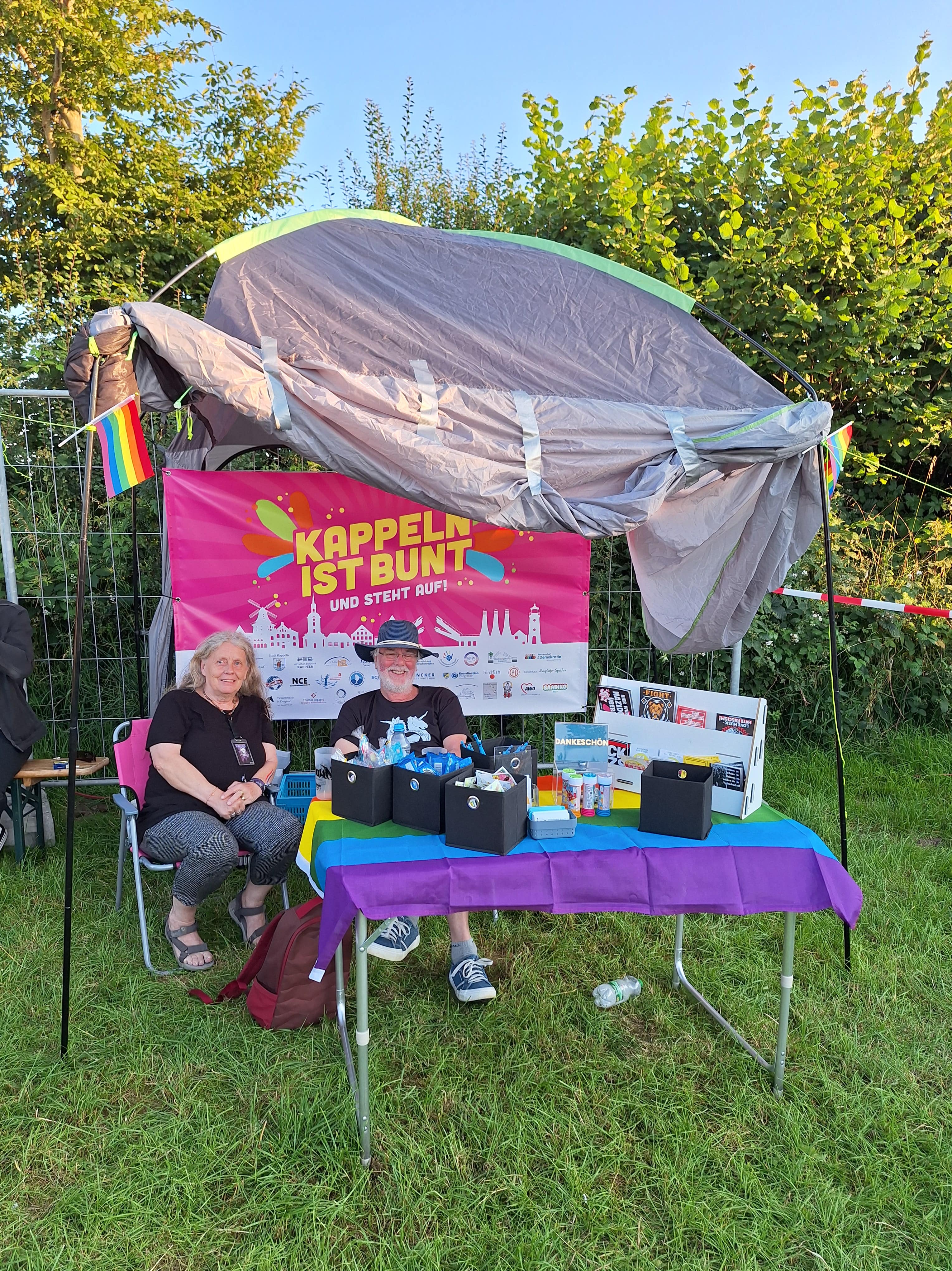 Ein Mann und eine Frau sitzen unter einem Sonnenschutz hinter einem Tisch und lächeln in die Kamera. Im Hintergrund hängt ein buntes Banner auf dem "Kappeln ist bunt und steht auf!" steht.
Der Stand ist mit Regenbogenfahnen und Flaggen geschmückt.
Auf dem Tisch gibt es Sticker, Süßigkeiten, Infokärtchen und eine Spendenbox.