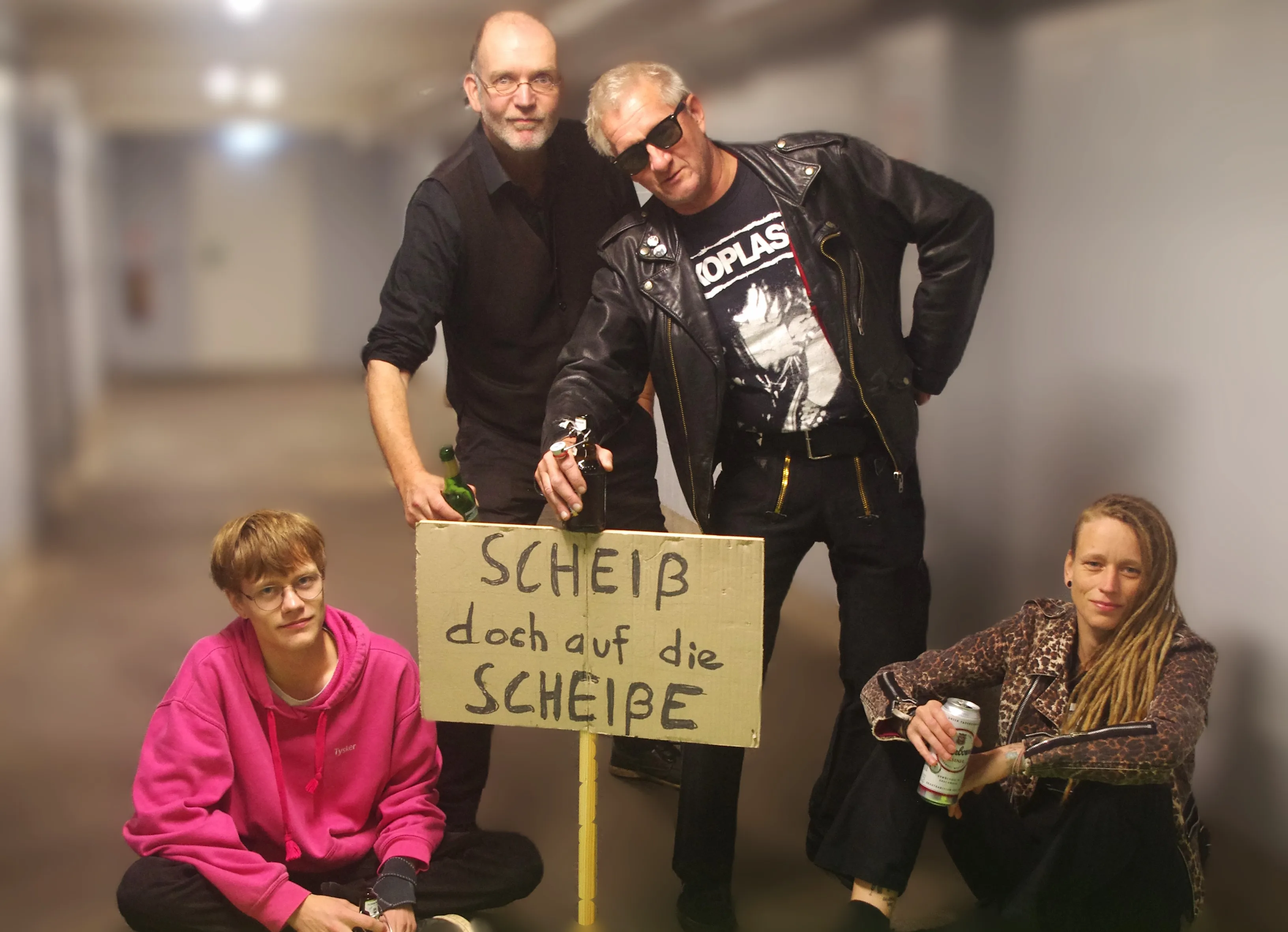 Vier Leute in einem Flur. Zwei stehen, zwei sitzen. Die stehenden lehnen auf einem Plakat auf dem "Scheiß doch auf die Scheiße" steht.