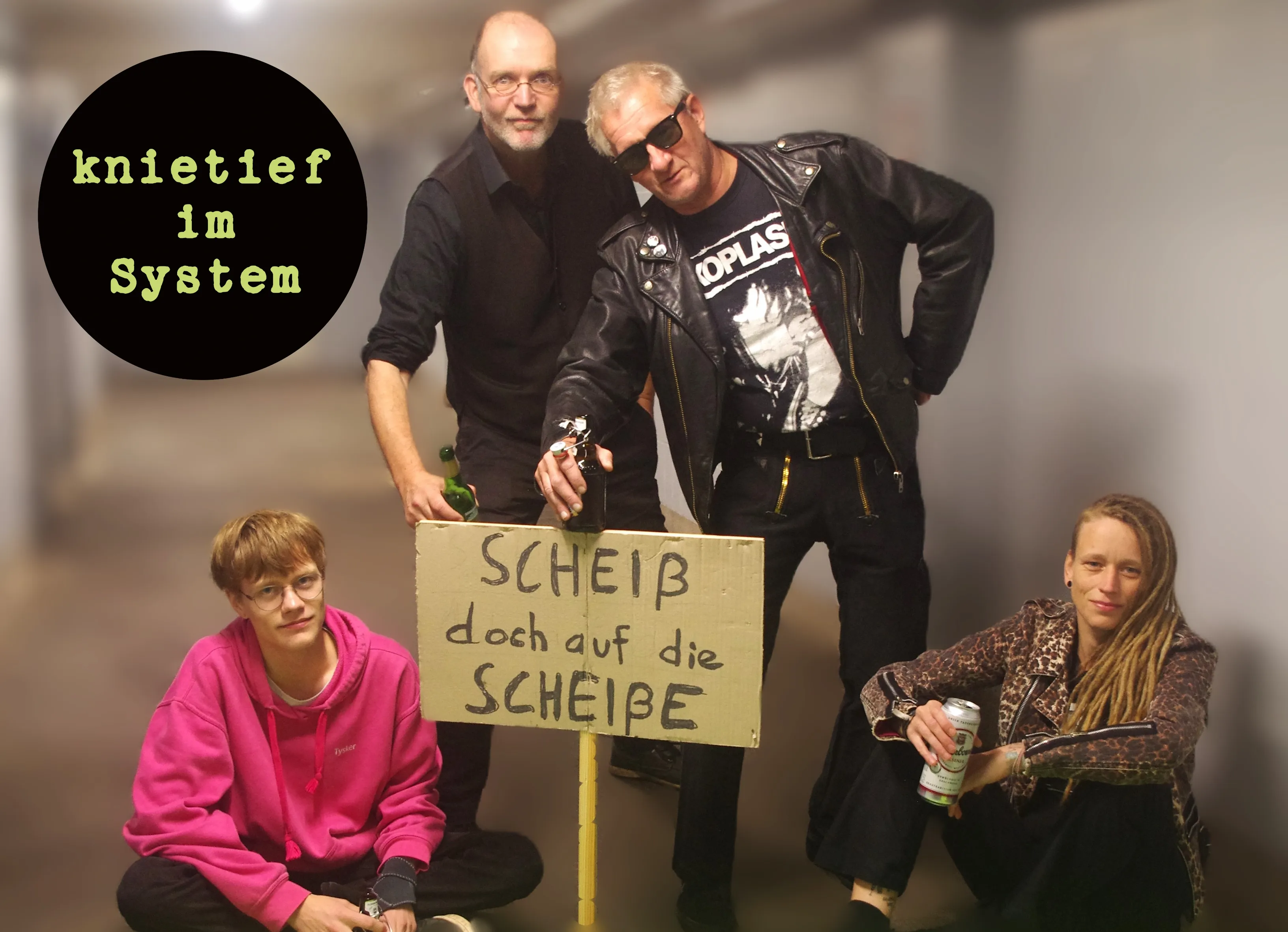Vier Leute in einem Flur. Zwei stehen, zwei sitzen. Die stehenden lehnen auf einem Plakat auf dem "Scheiß doch auf die Scheiße" steht.
Oben links im Bild das Bandlogo.
