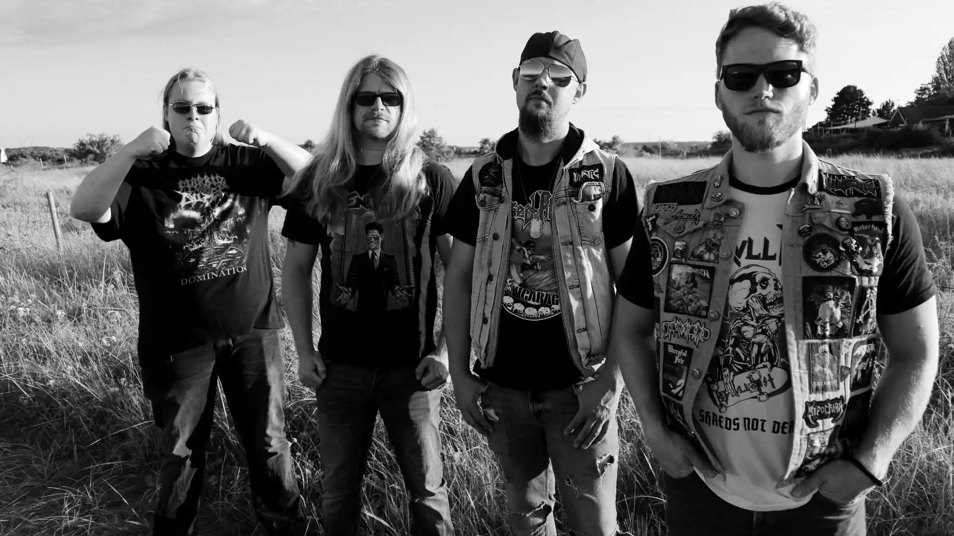 Vier Männer stehen nebeneinander auf einem Feldweg, hinter ihnen das Feld.
Sie tragen Bandshirts und Sonnenbrillen, zwei haben Metal-Kutten an.
Sie schauen neutral in die Kamera, der linke hat spaßig die Fäuste gehoben.