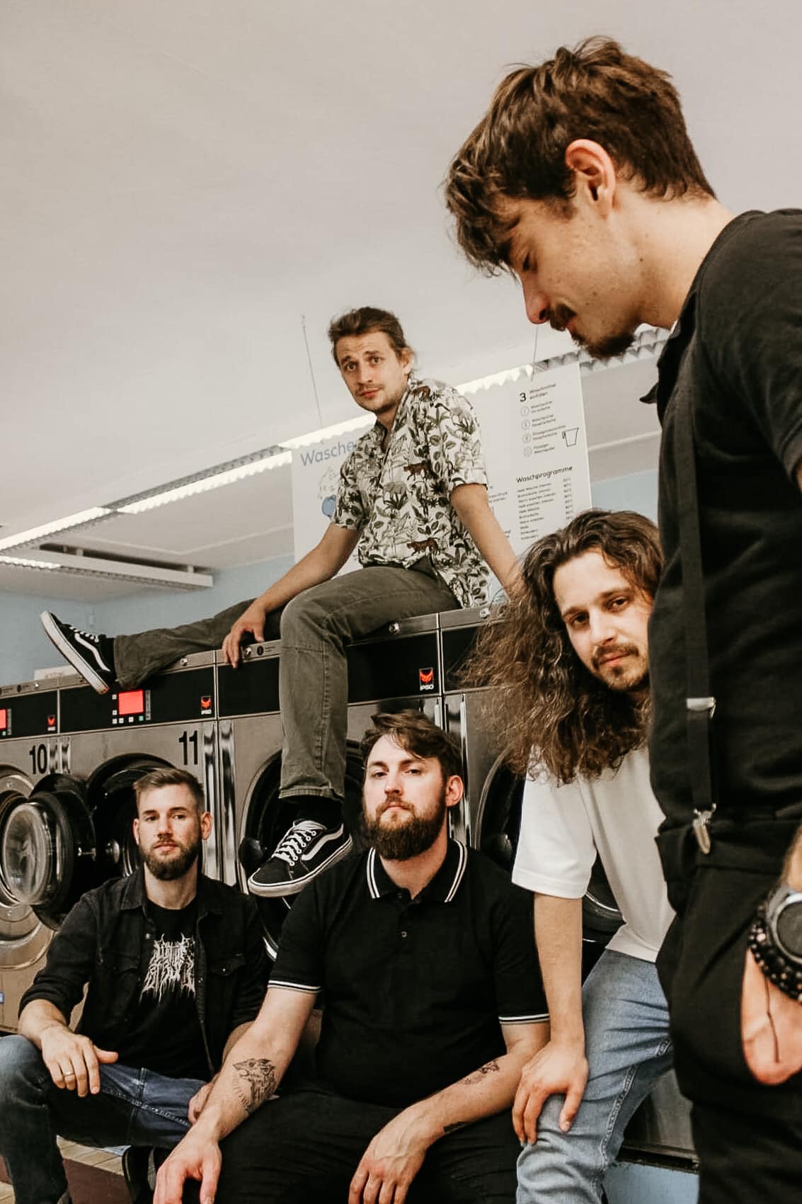 Bandfoto von Mass Rift.
Fünf Männer in einem Waschsalon.
Zwei hocken vor den großen Waschmaschinen, einer lehnt sich an einer steht und einer sitzt auf einer der Maschinen.