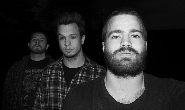 Bandphoto of Old Stone. Three guys stand diagonally behind each other in the dark and look into the camera. You can see them chest up. The image is black and white.