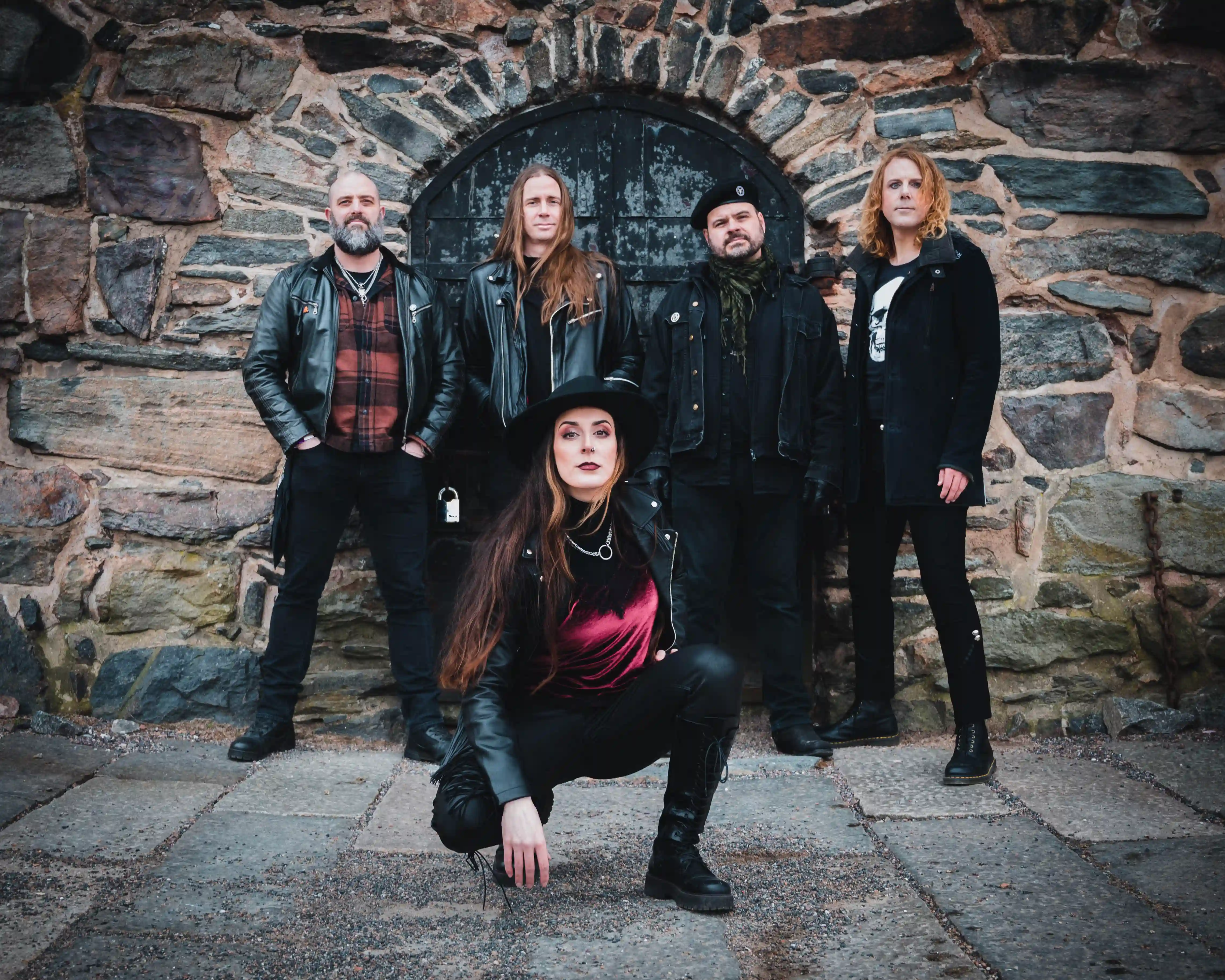 Bandfoto von Scarblade.
Vier Männer stehen  vor einem schwarzen Holztor, dass in eine grobe Steinmauer eingelassen ist.
Vor ihnen hockt die Frontsängerin. Alle sind schwarz gekleidet und schauen aufmerskam in die Kamera.