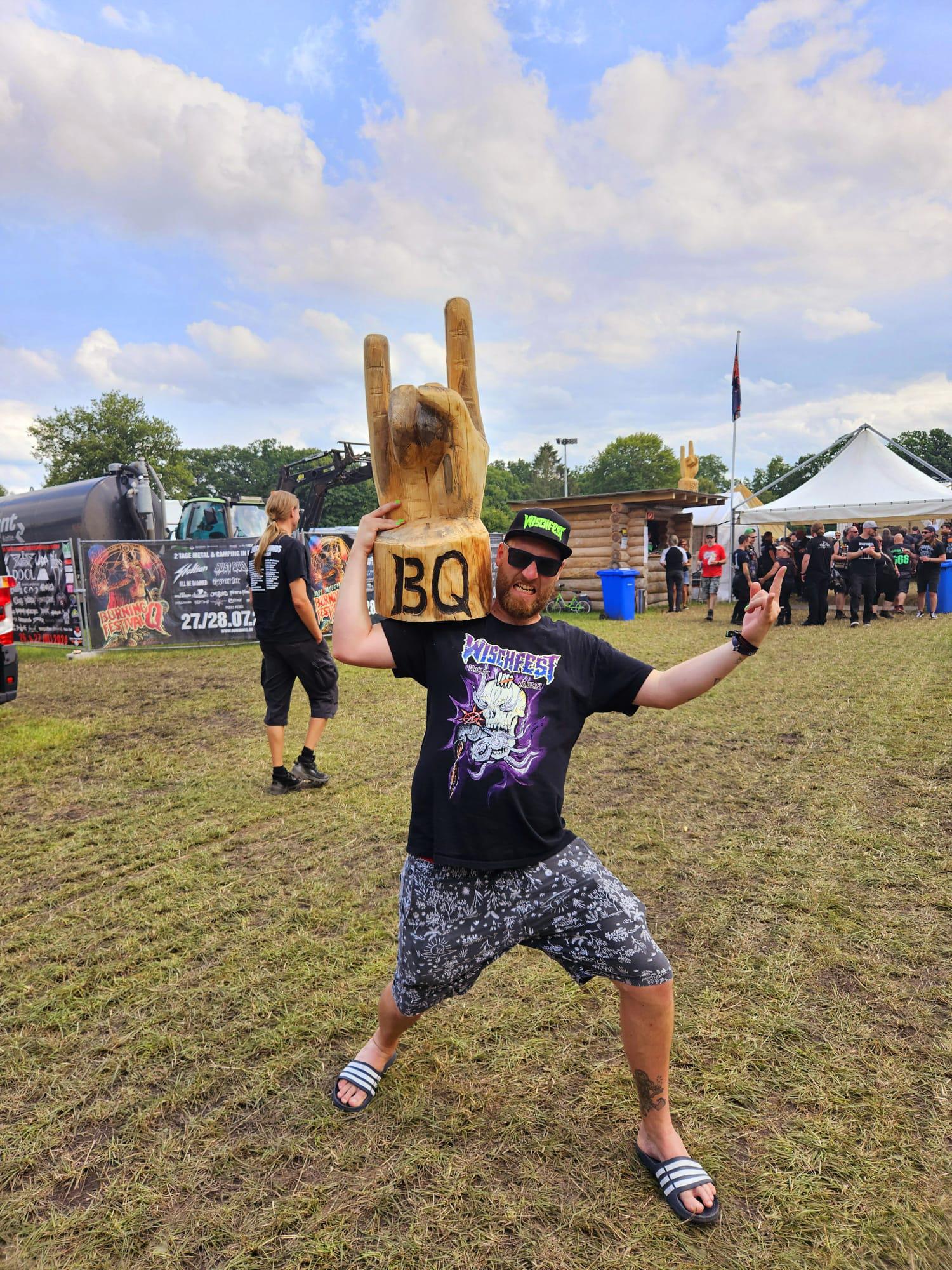 Ein Mann steht auf einem Festival mit Wischfest Cap und Wischfest T-Shirt und posiert mit einer großen hölzernen Pommesgabel auf der Schulter, auf der "BQ" für Burning Q (ein Festival) eingebrannt ist.