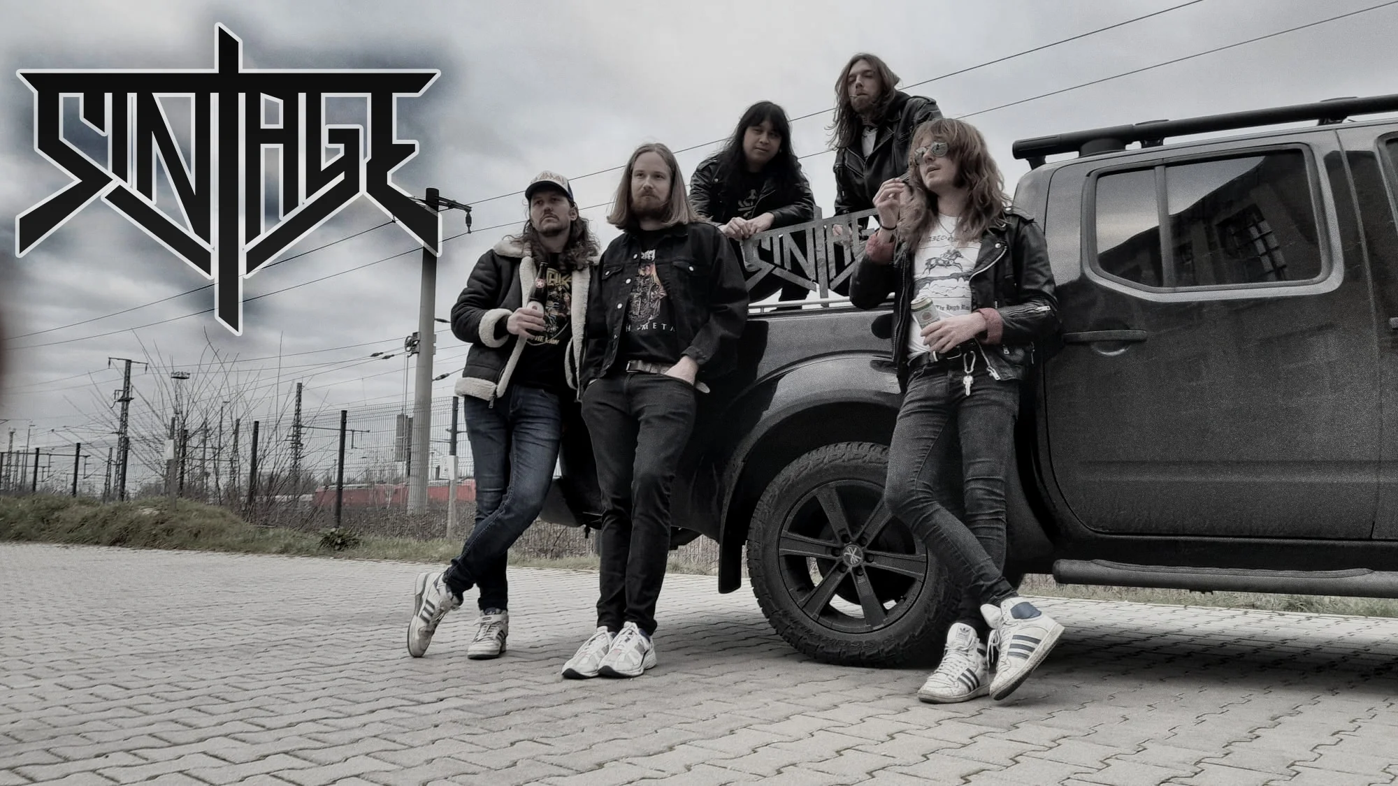 A dark pick up truck stands on paved floor.
Three men are leaning against it's loading space, two of them with beer cans in their hands.
Two other men are squatting at the loading space, one of them holding their bandlogo made of metal in his hand.
In the background are electricity pylons.
In the upper left corner is their bandlogo in black with transparent background.