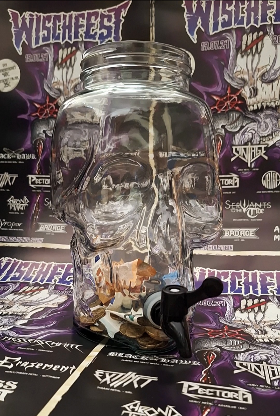 A photo of a glass skull with a tap in its mouth in front of some Wischfest 2024 posters.
The skull is about 30cm high.
Inside you can see various small change and a few banknotes.