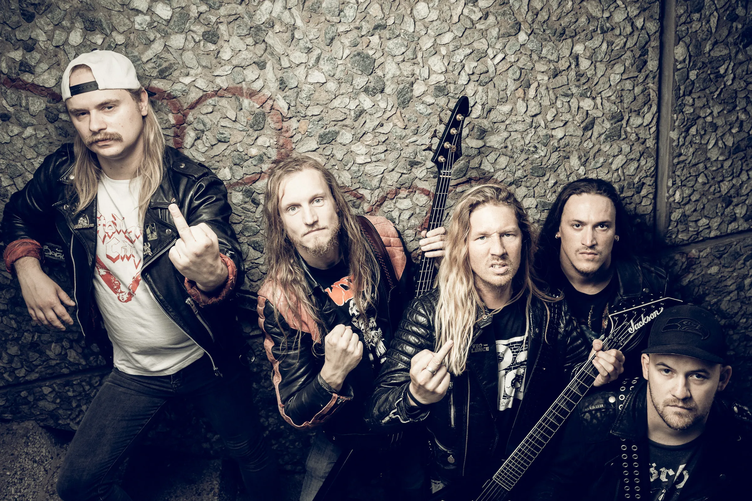 Fünf Männer stehen vor einer Steinwand und schauen rauf in die Kamera.
Einer hält einen Bass, einer eine Gitarre. Zwei zeigten Mittelfinger, einer eine Faust.