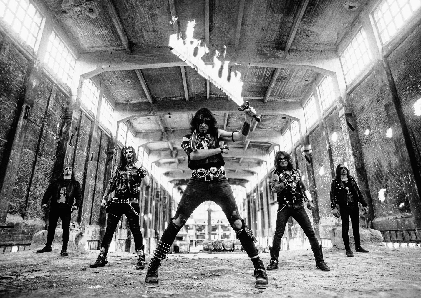 Five men stand in an empty warehouse and pose for the camera.
Their faces are painted black and white. The man in the front holds a flaming sword.
The image is in black and white.
